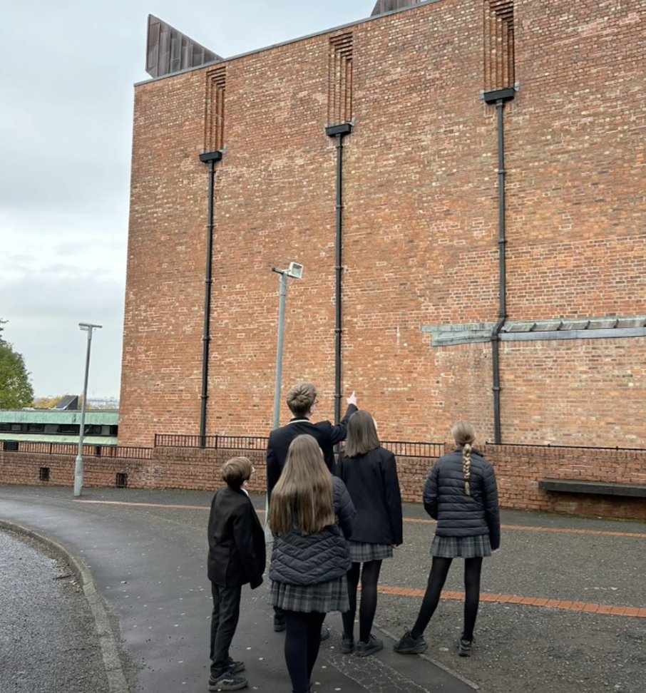 Pupils in school uniform looking up at a tall brick building while one points.