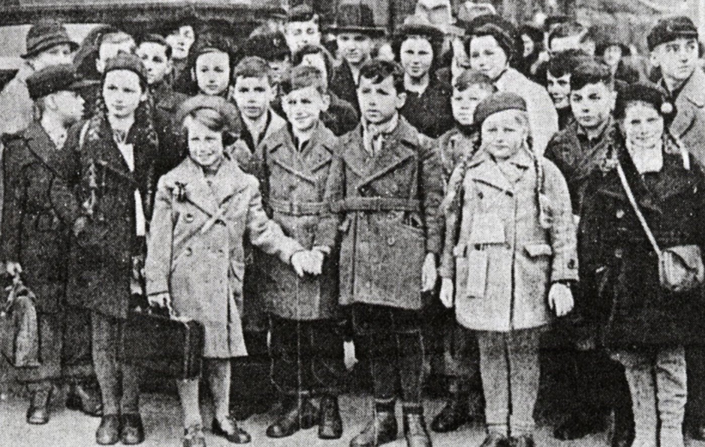 Black and white photograph of a large group of children in coats and hats, several holding suitcases.