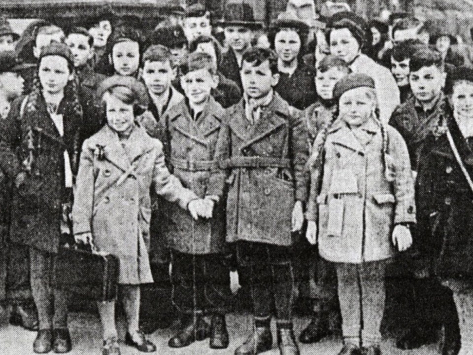 Black and white photograph of a large group of children in coats and hats, several holding suitcases, standing close together outdoors.
