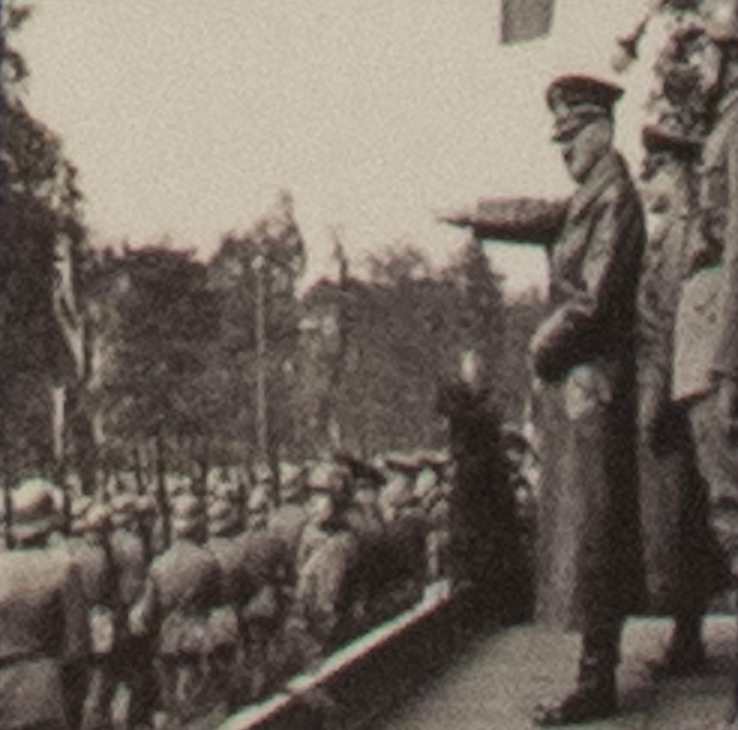 Sepia photograph of a uniformed man with his arm raised on a stand, watching rows of German soldiers march past.