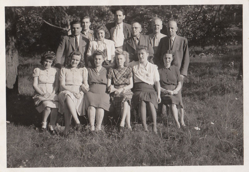 Black and white group photograph outdoors: six young women seated on the grass in front of seven standing men and women, taken in the 1940s.