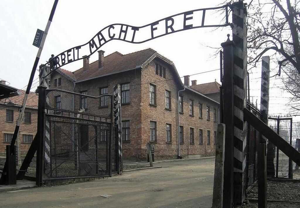 The entrance gate of Auschwitz with the iron words Arbeit macht frei above it, and brick barracks and barbed wire behind.