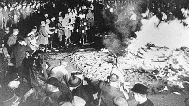 Black and white photograph of a crowd gathered around a large fire of burning books at night.